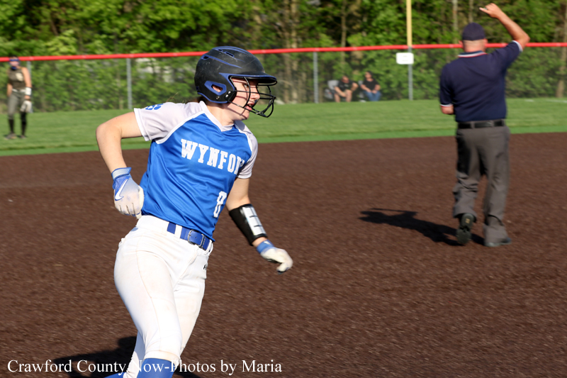 Baseball/softball player in a blue Wynford jersey runs on the infield dirt, wearing a helmet and white pants; umpire and spectators are visible in the background.