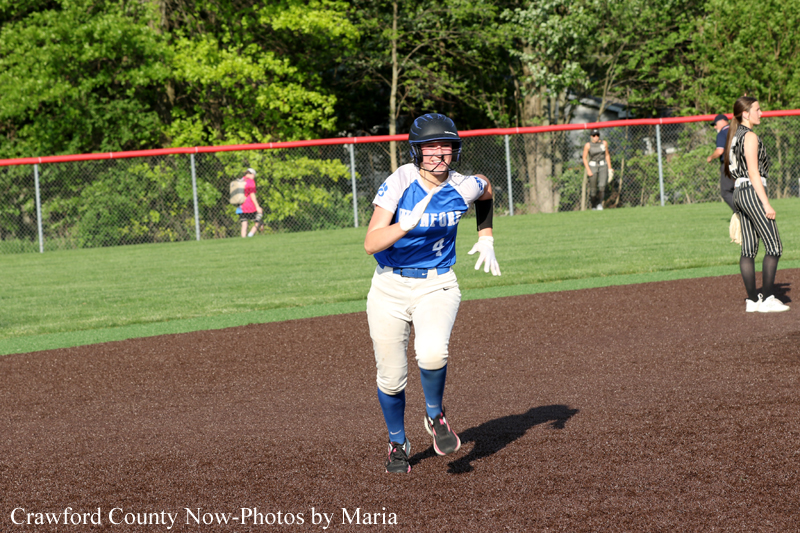 Female softball player in blue jersey runs on the infield dirt during a game, helmet on and glove ready, with fence and grassy outfield behind.
