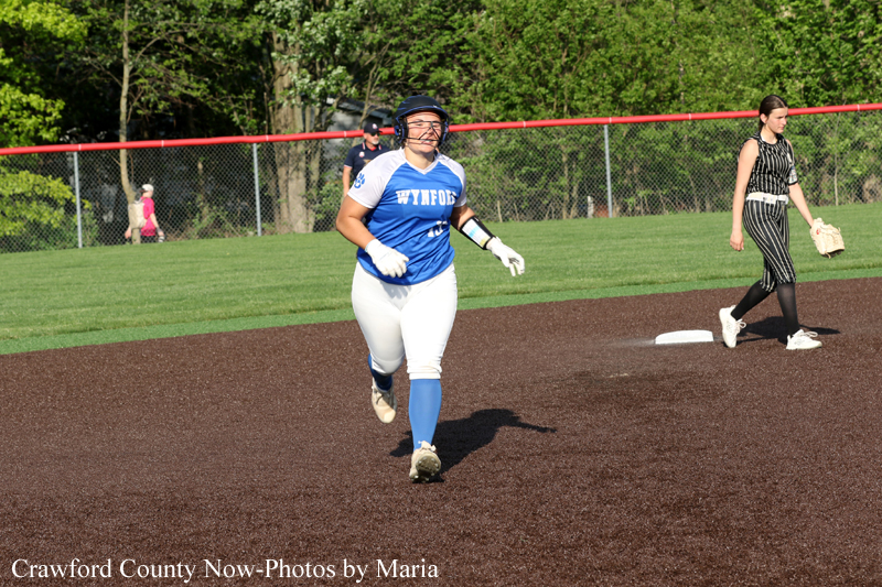 Softball player in a blue and white uniform runs on the infield dirt during a game, wearing a helmet and gloves; another player in a striped dress stands near base with a chain-link fence and trees in the background.