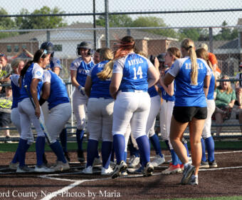 Girls' softball team in blue uniforms huddling on the infield during a game, with spectators in the stands behind a chain-link fence.
