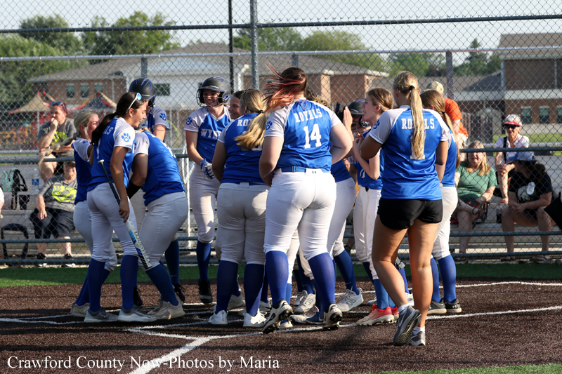Girls' softball team in blue uniforms huddling on the infield during a game, with spectators in the stands behind a chain-link fence.
