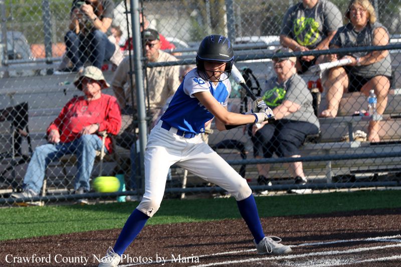 Baseball batter in blue and white uniform swings at a pitch during a game, with spectators in bleachers behind a chain-link fence.