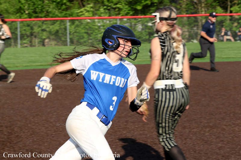 Young female softball player in a blue and white uniform (#3) runs on a dirt infield, wearing a helmet, during a game with a striped-clad opponent nearby.