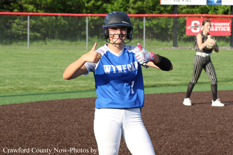 Softball player in a blue uniform and helmet giving two thumbs up on the field; another fielder in striped pants stands in the background.