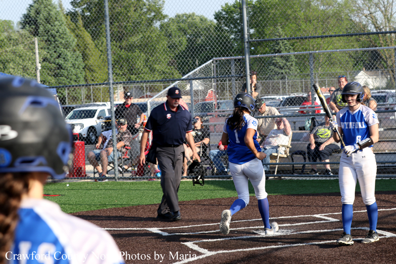 Blue-uniform softball players near home plate with an umpire watching as a runner approaches the plate.