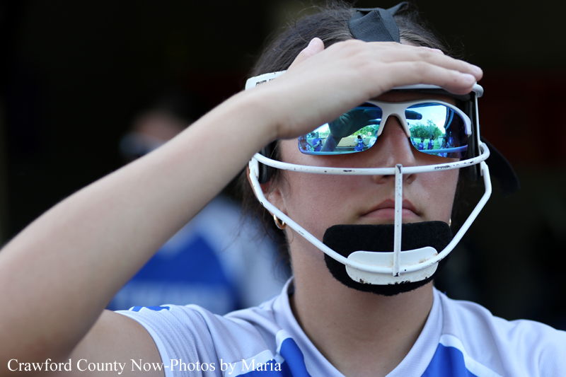 Female athlete wearing a white protective helmet with a black chin guard, shielding her eyes with a hand in a sunny field.