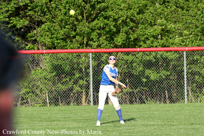 Female softball fielder in blue and white prepares to catch a rising ball on a grassy field.