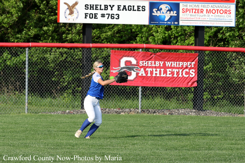 Young female softball fielder in a blue jersey and white pants reaches to catch a ball on a sunny grassy field, with red outfield fence and banners in the background.