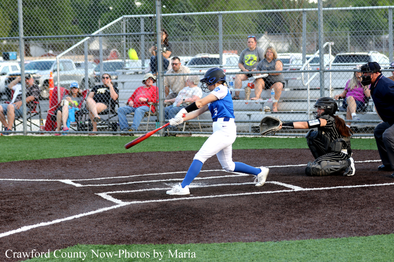 Young softball batter in a blue and white uniform swings at a pitch while catcher and umpire leer on behind home plate, with spectators in the background.