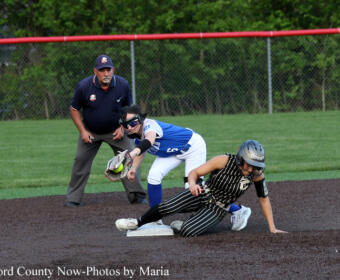 Softball baserunner slides into a base as a fielder in blue guards the bag; an umpire watches nearby on a dirt infield.