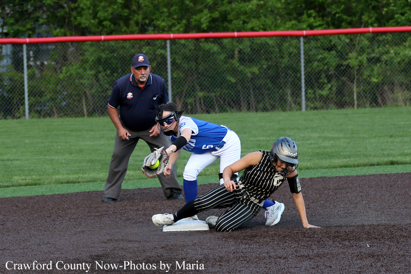 Softball baserunner slides into a base as a fielder in blue guards the bag; an umpire watches nearby on a dirt infield.