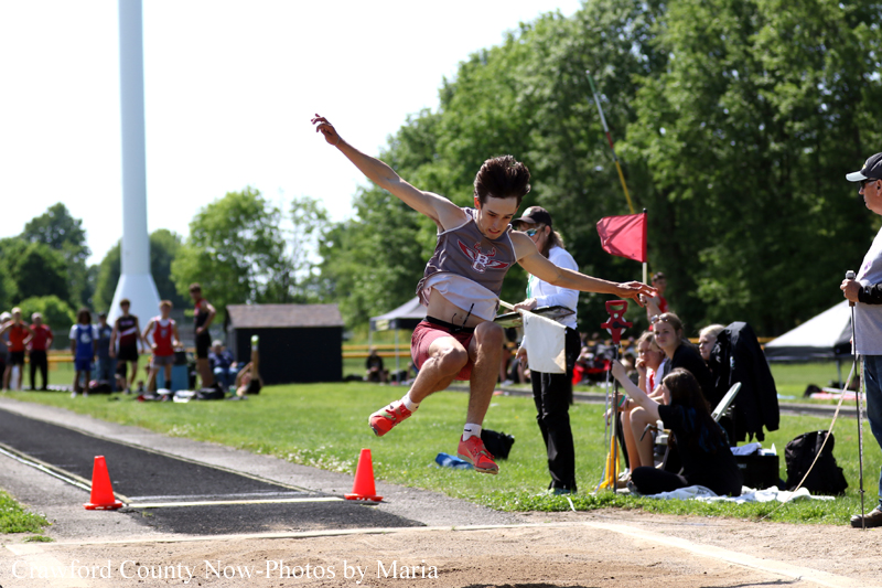 Female athlete in mid-air during a long jump at an outdoor track meet, landing area with orange cones and spectators in the background.