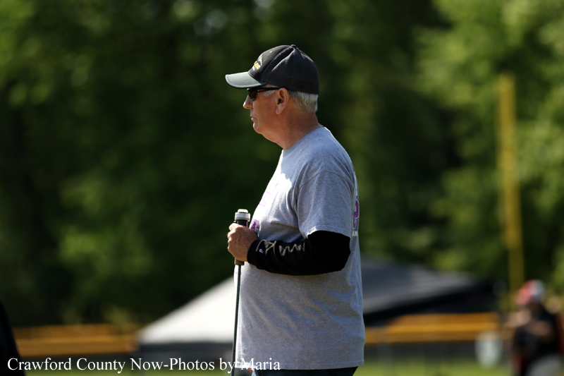 Man standing in profile outdoors, wearing a cap and sunglasses, holding a mic or baton in one hand.