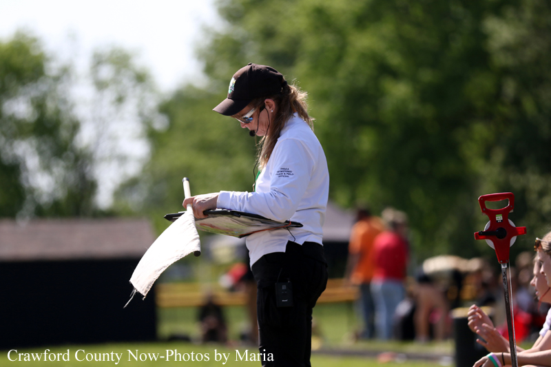 Female official wearing a white jacket and cap, jotting notes on a clipboard at an outdoor sports field.