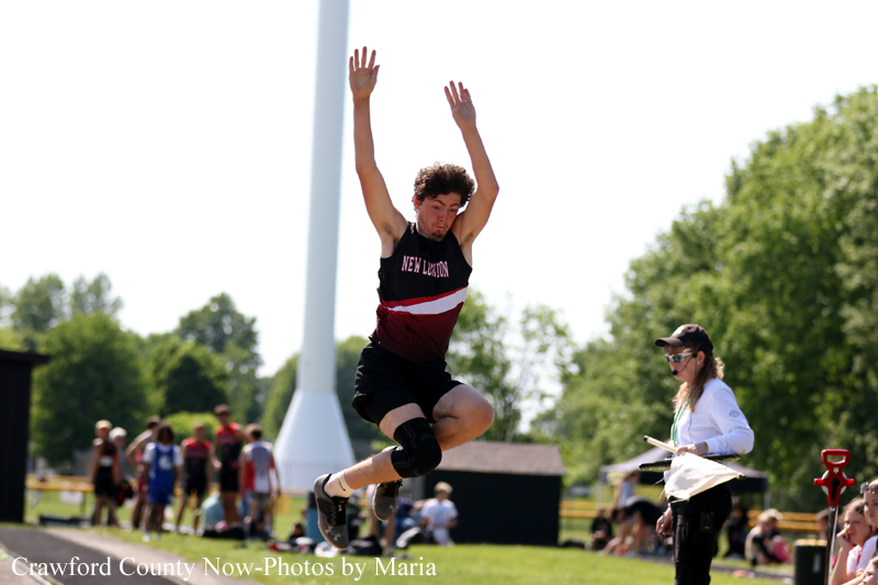 Male long jumper in a black and red uniform leaps mid-air on a sunny track field, with spectators in the background and an official watching nearby.