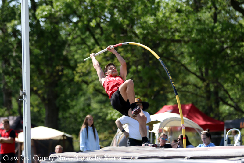 Pole vaulter in red shirt clears the bar during an outdoor track meet, mid-air with curved pole.