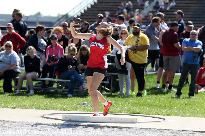 Female shot-put athlete in red top and black shorts performing a throw in the circle while a crowd watches from the stands behind her.