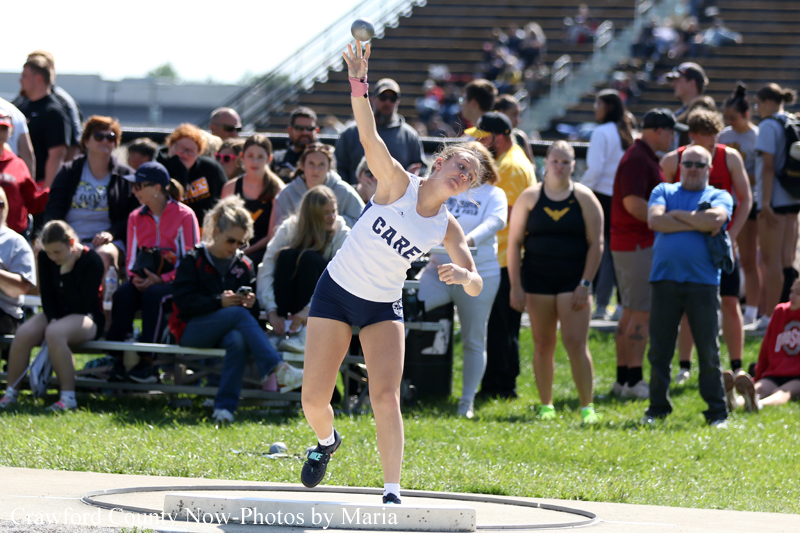 Female shot-put athlete in a white top and navy shorts throws the shot put from a circular throwing area, with a crowd watching in the background.