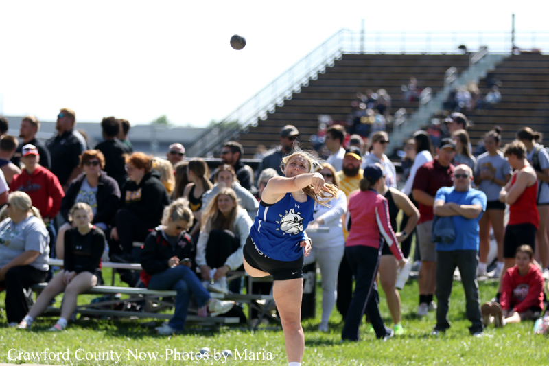 Female athlete in a blue jersey mid-throw of a shot put as a crowd watches from the bleachers behind her on a sunny field.