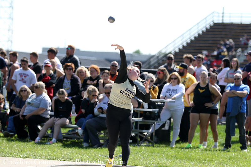 Female discus thrower in a beige uniform releasing a discus as a crowd watches at a track and field meet.