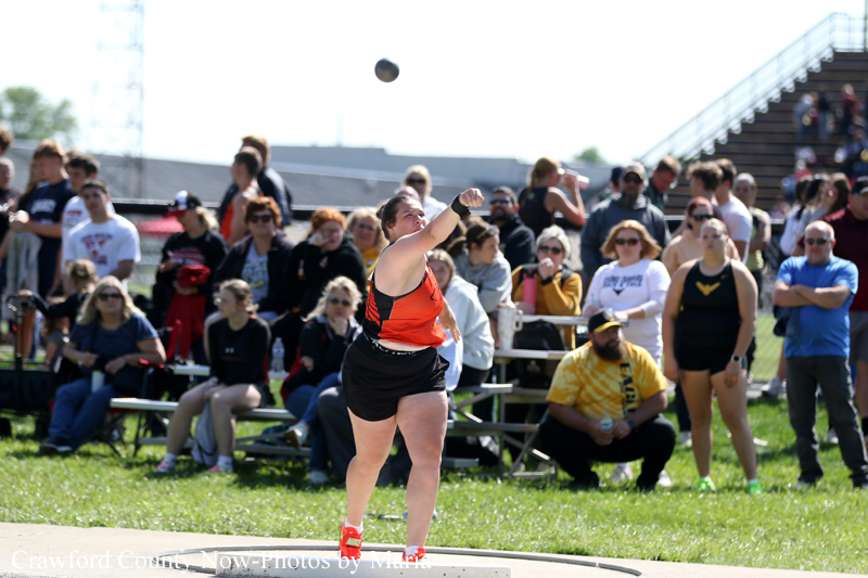 Female athlete in red top and black shorts throwing a shot put during a sunny outdoor event as spectators watch from bleachers and grass field behind her.