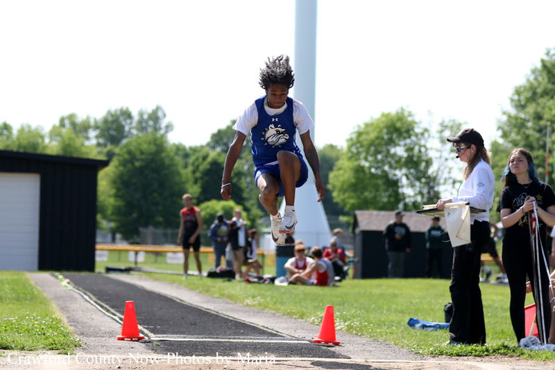 Young athlete in blue uniform mid-jump over a track during a field event, with cones and spectators watching in the background.