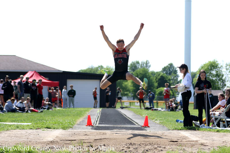 A male long jumper in a black and red uniform leaps mid-air along the track toward the sand pit, with spectators and officials watching in the background.