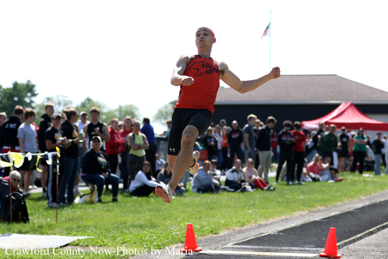 Male athlete in a red top mid-air leaping during a track and field event, with a crowd watching on a grassy field behind him.