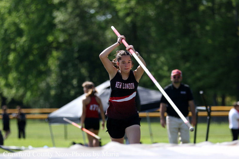Female pole vaulter in a New London uniform sprints with a pole during an outdoor track meet, with officials and tents in the background.