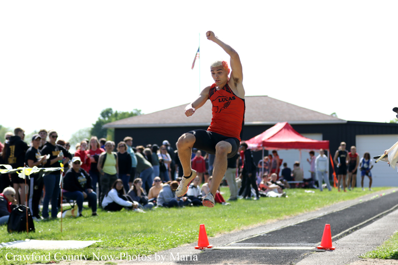 Male athlete in an orange singlet is mid-air performing a long jump, with a crowd watching in the background.