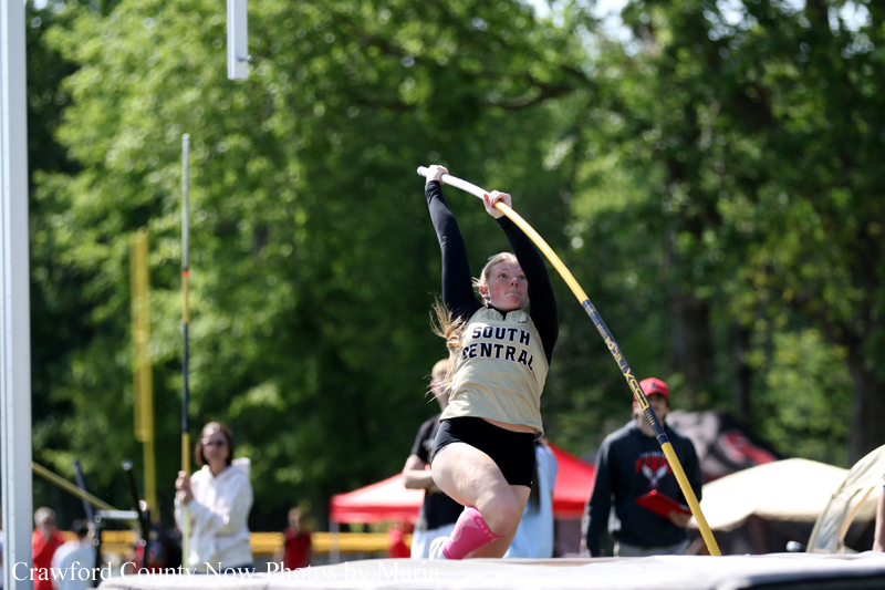 Female pole vaulter mid-air during an outdoor track meet, wearing a beige SOUTH CENTRAL shirt and black shorts.