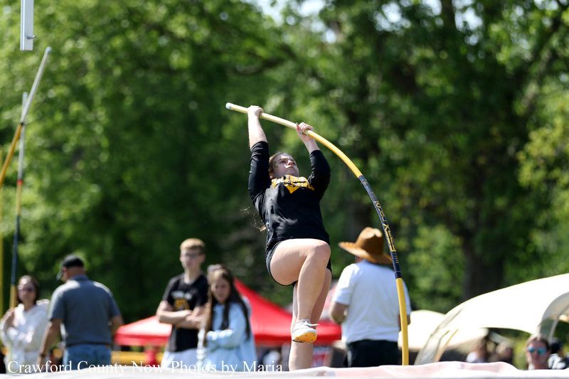 Pole vaulter in black clothing clearing a yellow bar outdoors as spectators look on from the field.