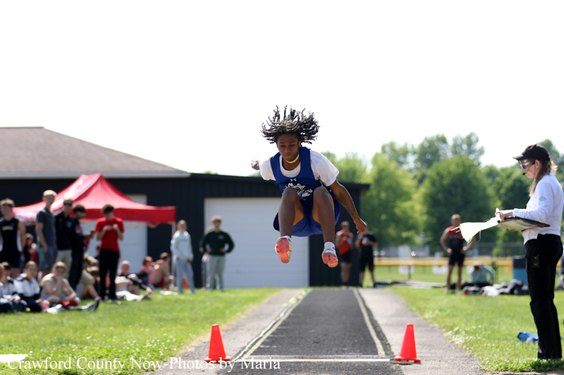 Young athlete mid-air clearing a hurdle on a track, with spectators and a coach nearby under a bright sky.