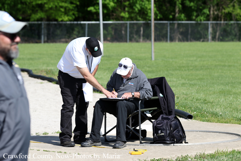 Two men outdoors by a fence; one sits at a folding chair filling a clipboard while the other stands beside him, offering help.