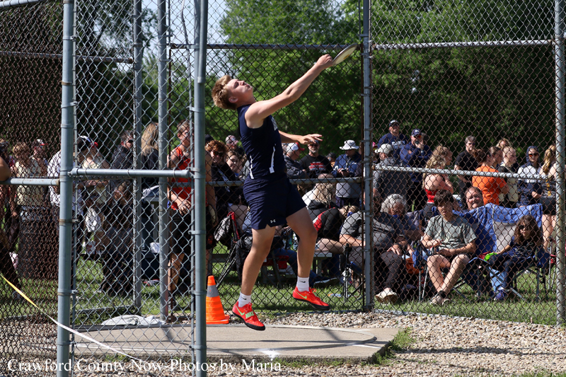 Male track athlete in navy attire releases a discus from a throwing circle inside a chain-link cage as spectators watch behind him.