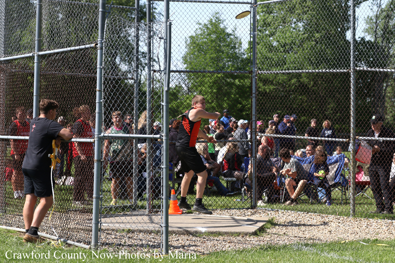 Teen throws a discus inside a chain-link fenced throwing cage as spectators watch nearby.