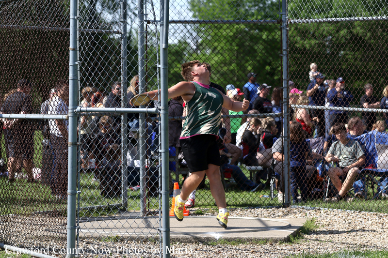 Athlete in a green sleeveless shirt and black shorts leaps inside a chain-link fenced throwing area as spectators watch nearby.