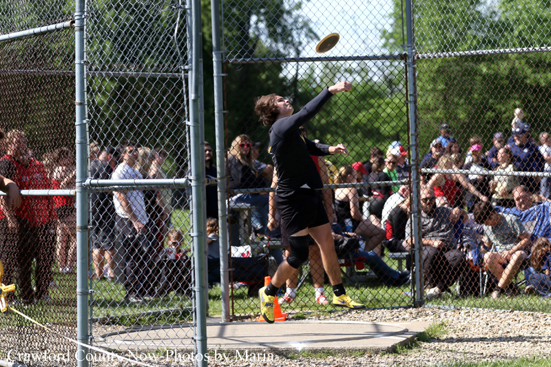 Athlete in black gear mid-throw of a discus inside a chain-link fenced throwing circle, with a crowd watching beyond the fence.