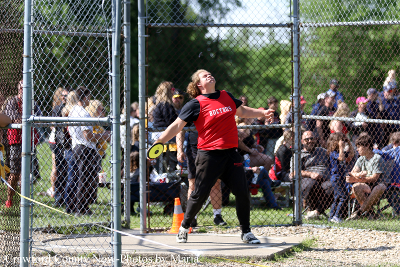 Athlete in a red shirt throwing a discus inside a chain-link fence; crowd watches nearby.