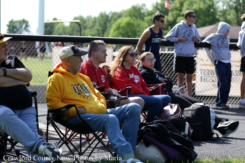 Group of spectators sitting in folding chairs along a track, watching an outdoor race on a sunny day.