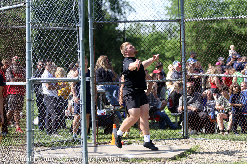 Athlete in a black shirt and shorts mid‑throw inside a chain‑link throwing cage, with spectators watching on a sunny day.