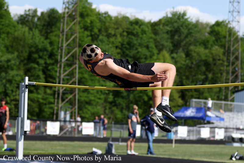 Male high jumper in black clears a yellow bar on an outdoor track, with spectators and green trees in the background.