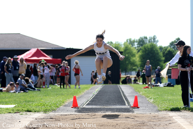 Female long jumper in a white uniform leaps through the air toward the sand pit as a crowd watches on a sunny day.