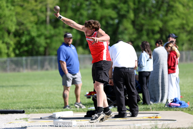 Athlete in a red jersey throws a shot put on a grassy field, with spectators watching in the background.