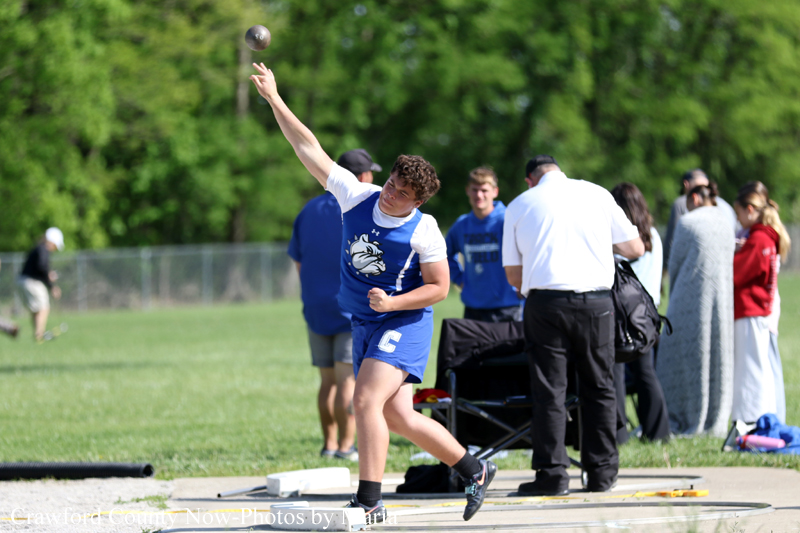 Male discus thrower in blue and white uniform releases a discus on a grassy field while teammates and spectators watch in the background.