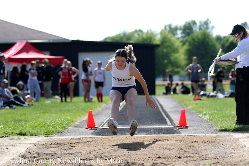 Female long jumper in mid-air during a track meet, about to land in the sand pit as spectators watch.
