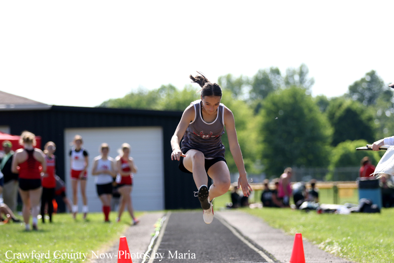 Female athlete in a gray tank top and dark shorts leaps mid-air on a track runway, with orange cones and spectators in the background on a sunny day.