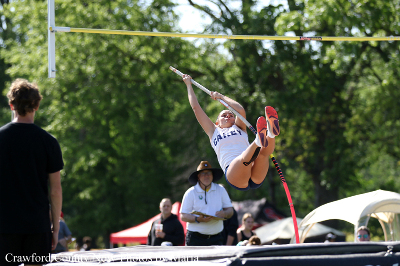 Pole vaulter in a white shirt clears the bar midair during a competition, with a judge and spectators in the background on a sunny day.