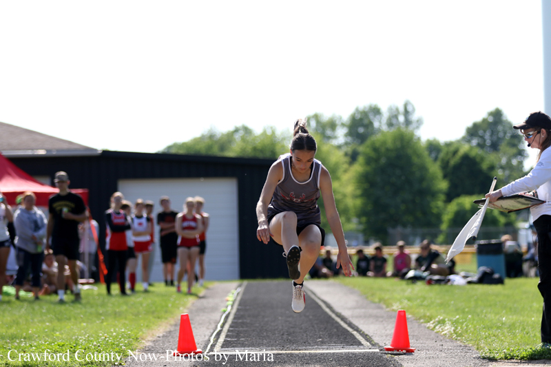 Female long jumper mid-air during a competition, landing pit visible ahead with cones and onlookers in the background outdoors.