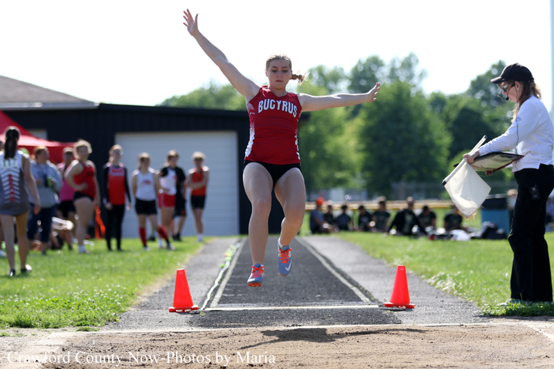 Female athlete in a red Bucyrus uniform leaps mid-air on a track long jump runway, with cones and spectators nearby.